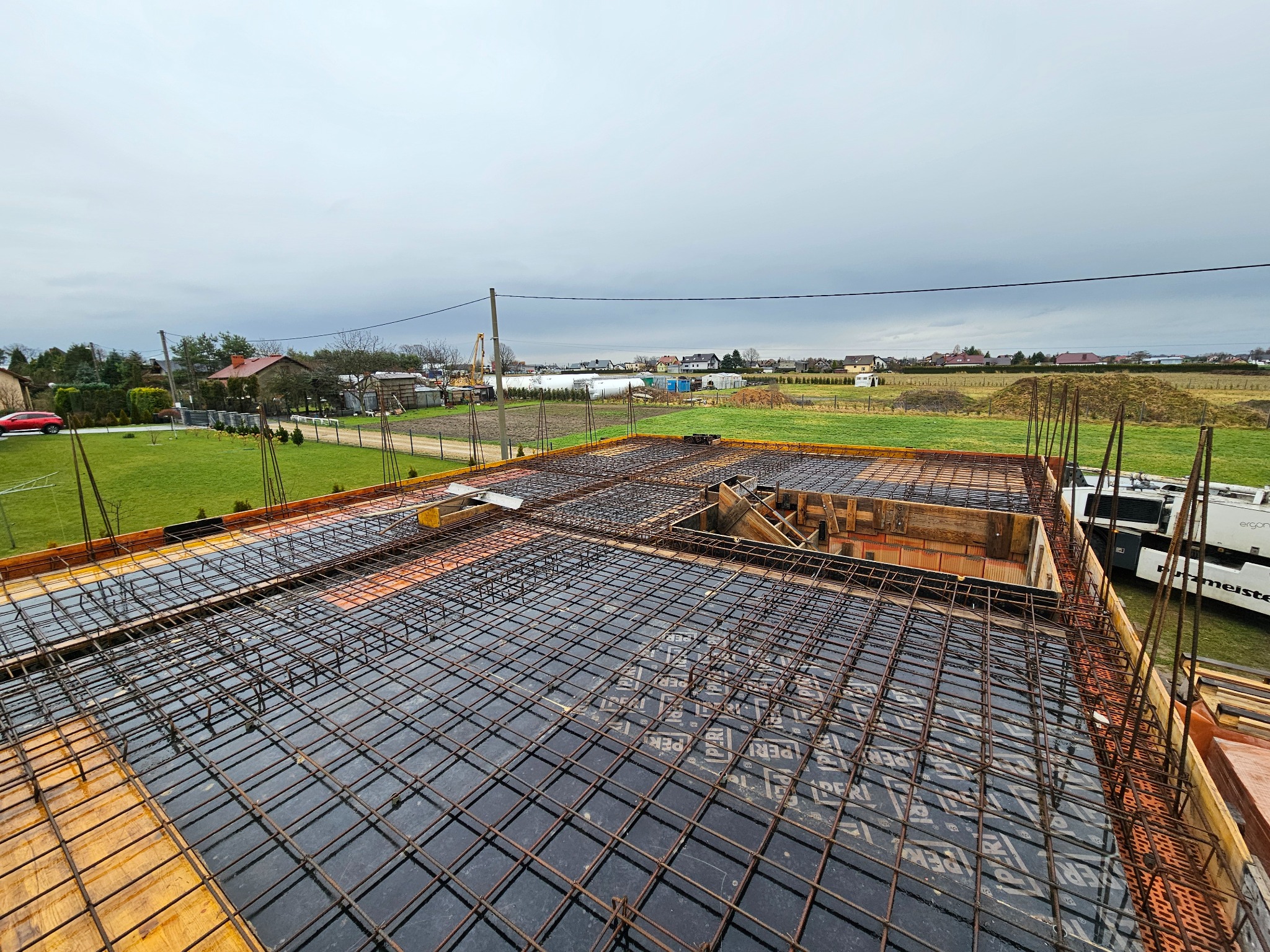 Zbrojenie płyty fundamentowej z widoczną siatką stalową i deskowaniem, przygotowane do zalania betonem. W tle zabudowa jednorodzinna i pochmurne niebo.