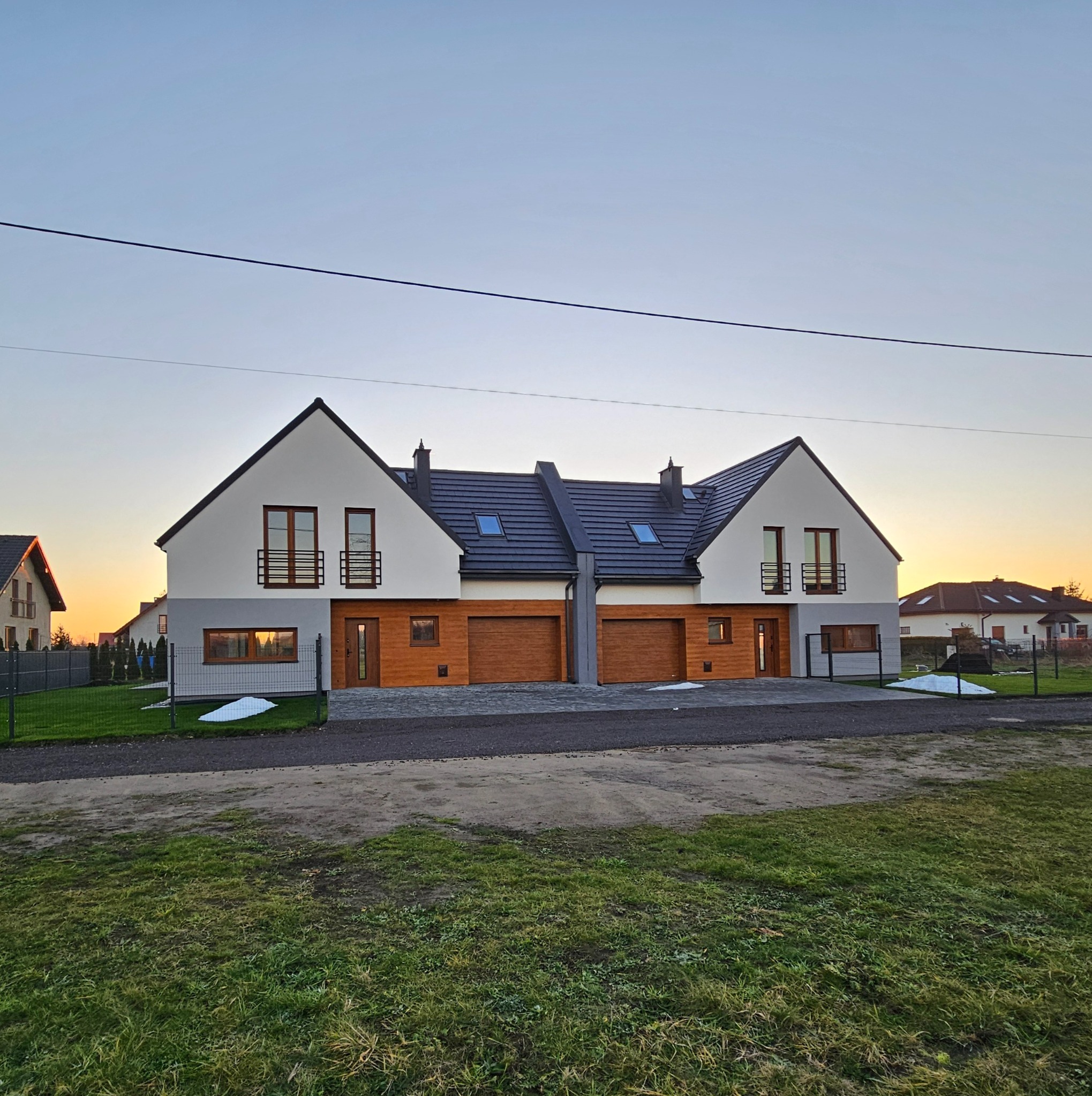 Nowoczesny bliźniak z drewnianą elewacją i garażami, sfotografowany o zmierzchu. Widoczne okna z balustradami i fragmenty ogrodzenia. Minimalistyczny design.