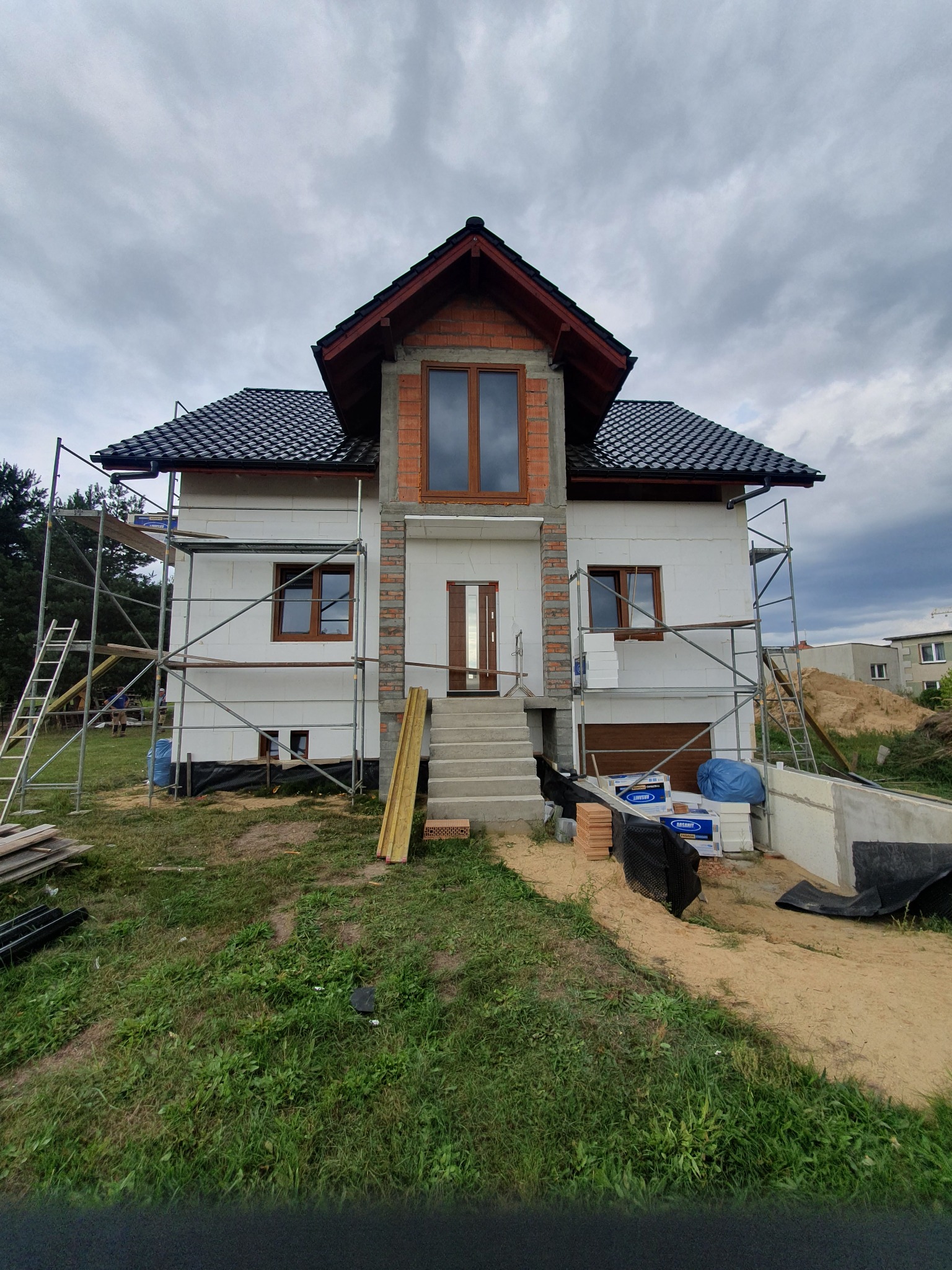 Niedokończony dom z białym styropianem na elewacji, ceglanym wykończeniem i rusztowaniami. Widoczne materiały budowlane i elementy konstrukcyjne.