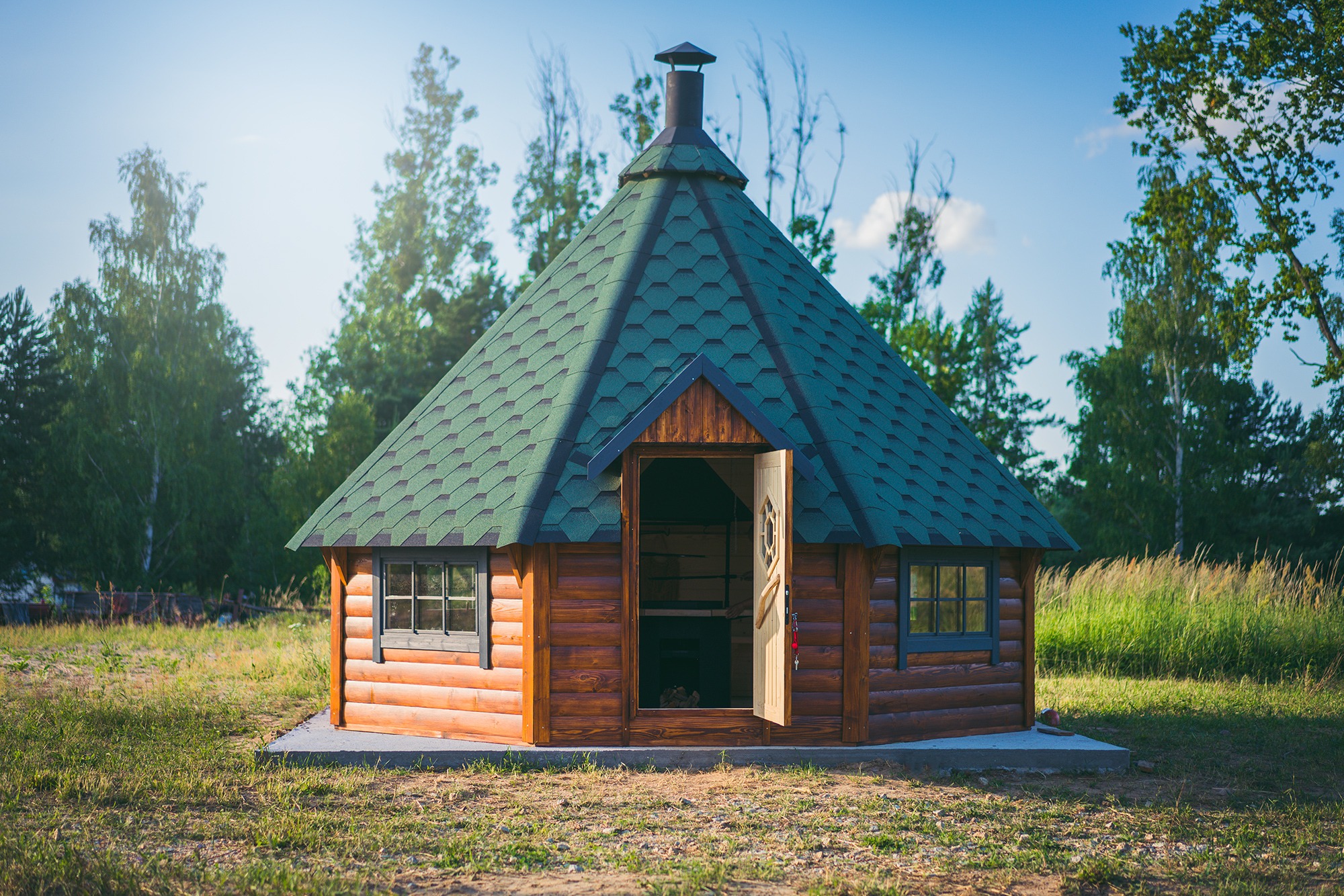 Drewniana altana z zielonym dachem krytym gontem bitumicznym, z otwartymi drzwiami, na betonowej wylewce, otoczona trawą i drzewami.