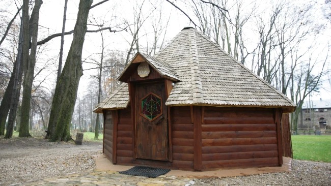 Drewniana sauna ogrodowa z dachem krytym gontem, sześciokątnym oknem z witrażem i okrągłym szyldem nad drzwiami, otoczona jesiennym krajobrazem.