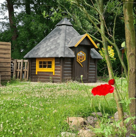 Drewniana altana w kształcie domku z żółtymi ramami okiennymi i drzwiami, ozdobiona motywem plastra miodu, otoczona zielenią i czerwonymi makami.
