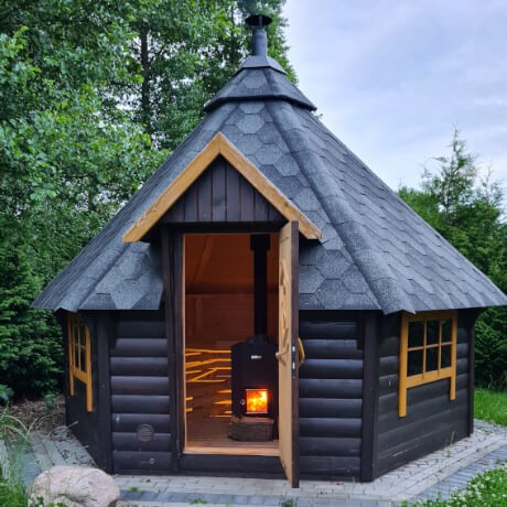 Ośmiokątna, ciemna, drewniana kabina z dachem krytym gontem, z otwartymi drzwiami, ukazującymi piec opalany drewnem wewnątrz, otoczona zielenią.