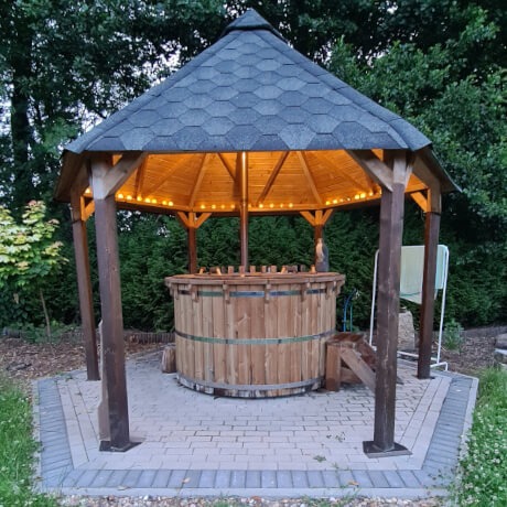 Drewniana balia umieszczona wewnątrz zadaszonej altany z oświetleniem dekoracyjnym, otoczonej zielenią.