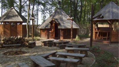 Trzy drewniane altany w lesie, jedna z kominem, otoczone ławkami z bali, stos drewna obok jednej z altan.