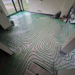 USŁUGI HYDRAULICZNE PRZEMYSŁAW ŚWIĄĆ - Zielone rurki ogrzewania podłogowego ułożone spiralnie na siatce w surowym wnętrzu, widoczne materiały budowlane i okno.