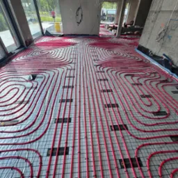 Czerwone rury ogrzewania podłogowego ułożone spiralnie na szarej macie w jasnym pomieszczeniu z betonową ścianą. Widoczne okna i osoba w oddali.