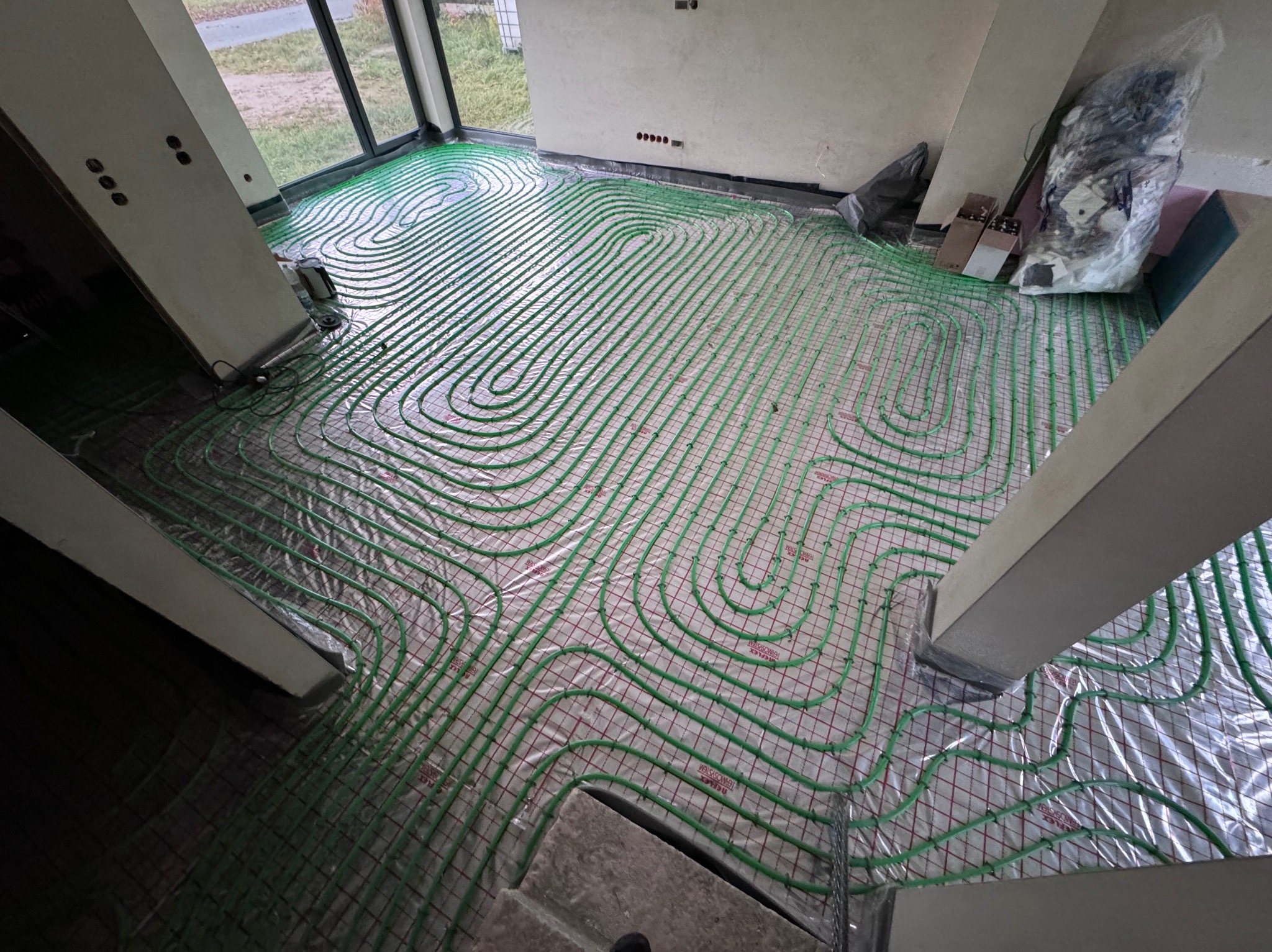 Zielone rurki ogrzewania podłogowego ułożone spiralnie na siatce w surowym wnętrzu, widoczne materiały budowlane i okno.