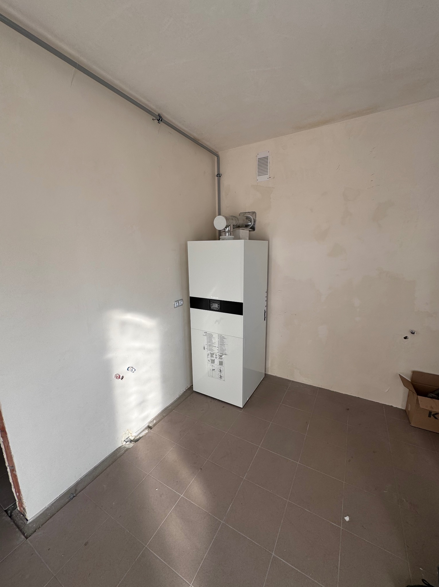 Biały piec centralnego ogrzewania z czarnym panelem, zamontowany w surowym pomieszczeniu z beżowymi ścianami i brązową podłogą z płytek, widoczne rury instalacyjne i karton po prawej stronie.