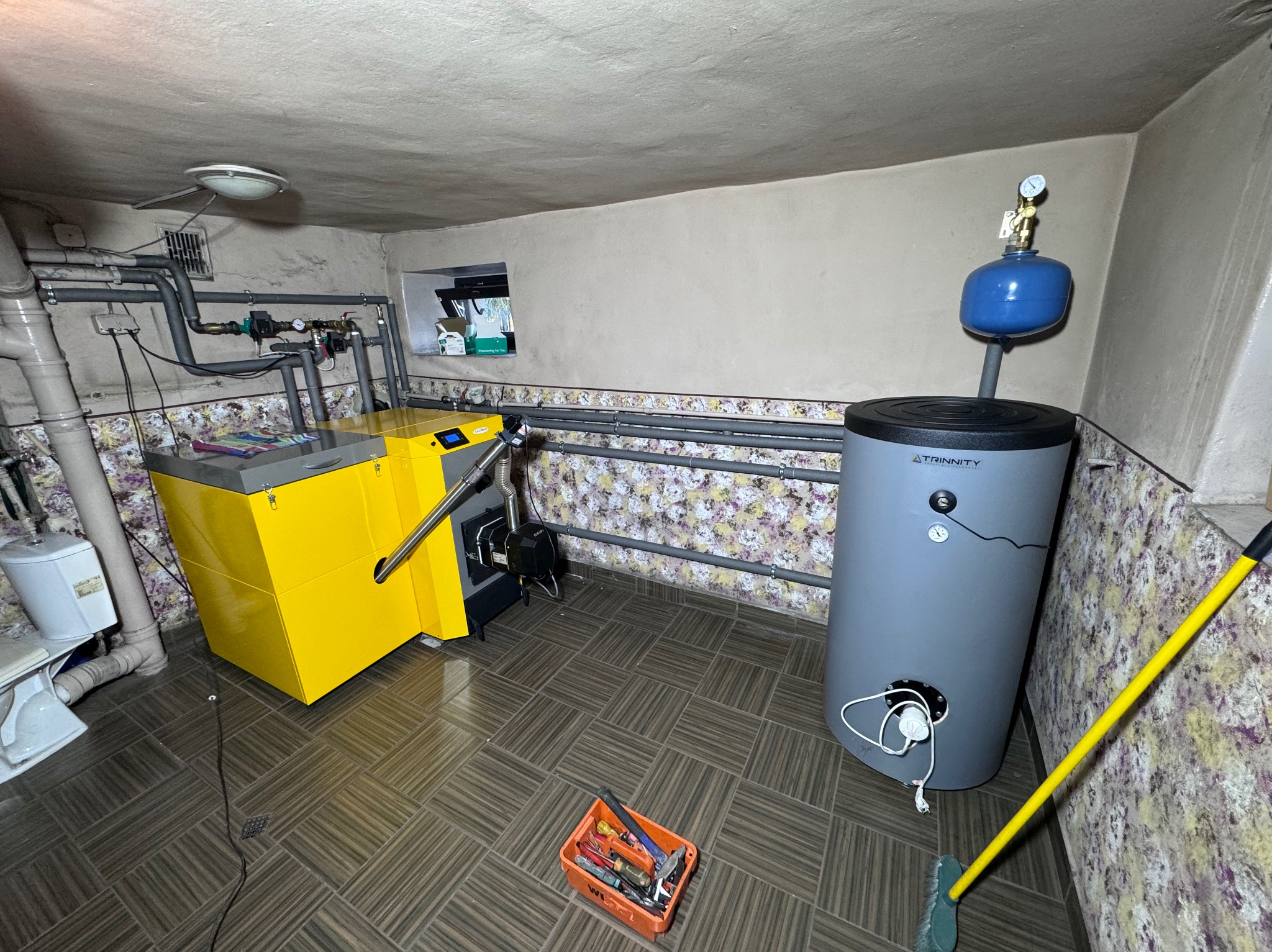 Kotłownia z piecem na ekogroszek, zbiornikiem buforowym, instalacją hydrauliczną i skrzynką z narzędziami na podłodze z płytek imitujących drewno.