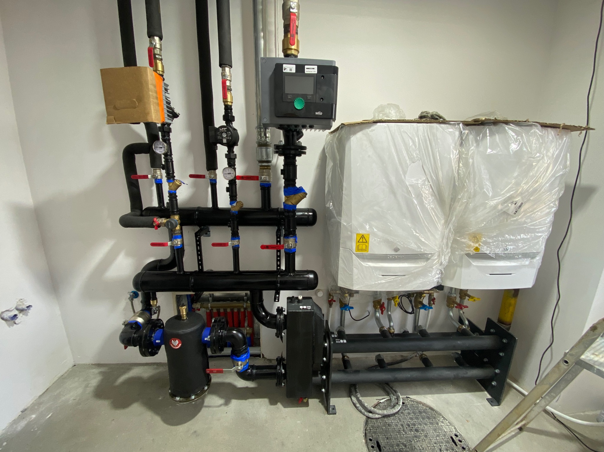 Złożona instalacja grzewcza z dwoma kotłami w folii ochronnej, widoczne rury, zawory, sterownik i elementy montażowe, na podłodze właz kanalizacyjny.