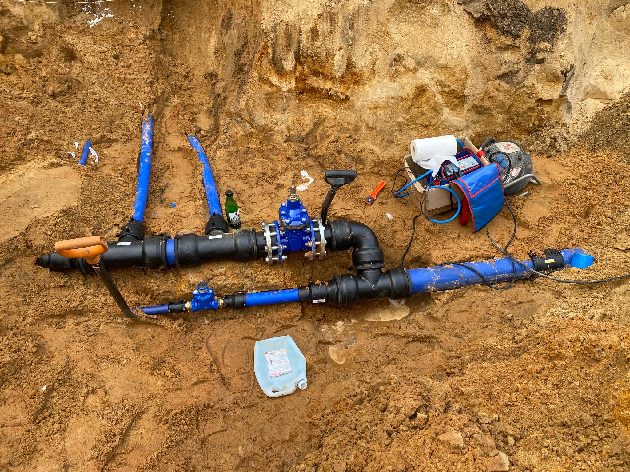 Wykop z widocznymi, częściowo ułożonymi rurami wodociągowymi w Częstochowie, z narzędziami i akcesoriami obok. Widoczne zawory, złączki i butelka.