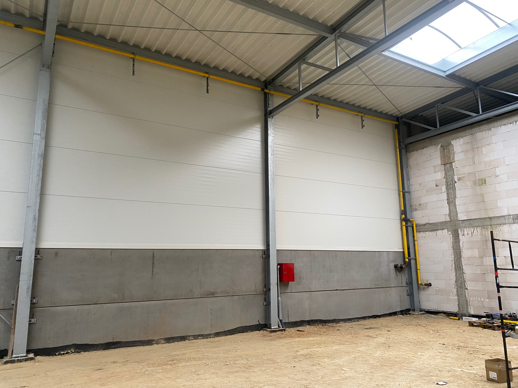 Wnętrze hali produkcyjnej w trakcie budowy, widoczne ściany z paneli warstwowych, instalacja gazowa w żółtej rurze, surowy betonowy cokół i fragment ściany z bloczków betonowych.