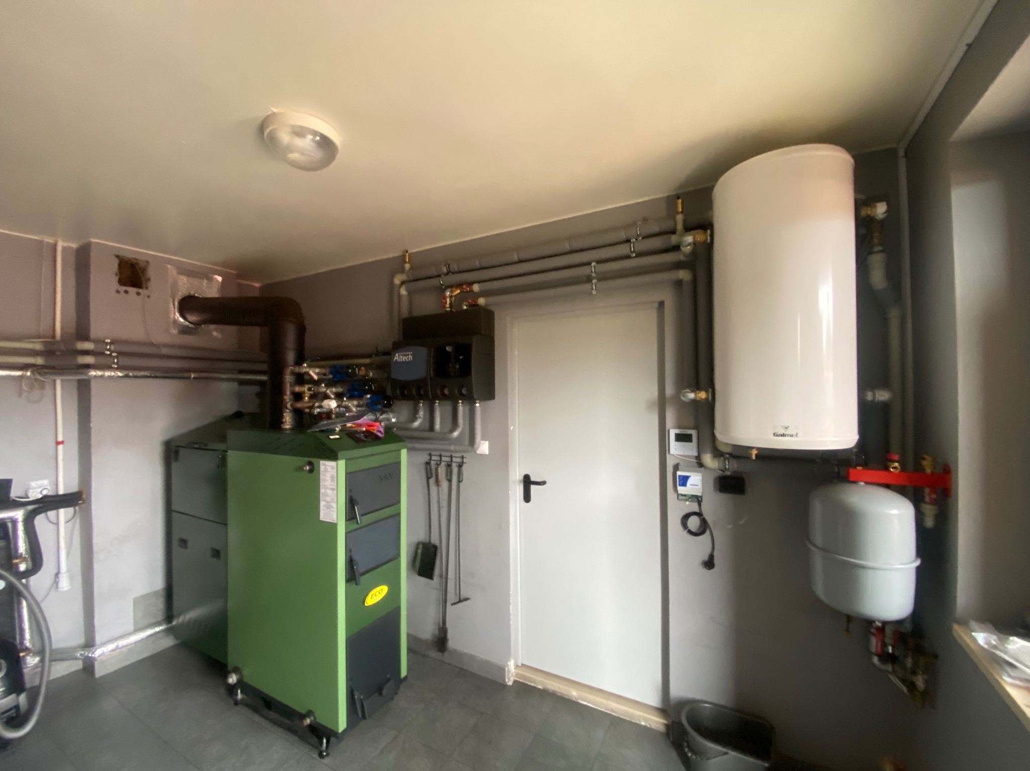Kotłownia z zielonym kotłem na paliwo stałe, zasobnikiem ciepłej wody i orurowaniem, widok na zainstalowany system grzewczy w pomieszczeniu z szarymi ścianami.