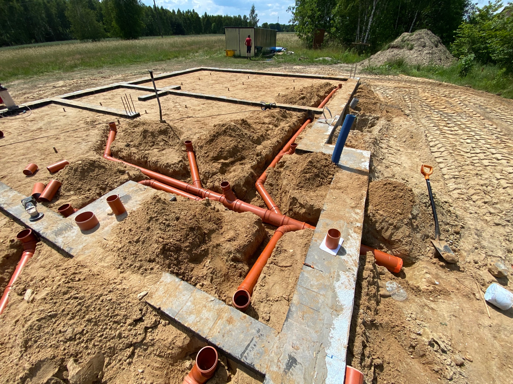 Instalacja kanalizacyjna z rur PVC w wykopie fundamentowym, widoczne połączenia i kształtki, ziemia zasypana częściowo, w tle zarys fundamentów i osoba przy szopie.