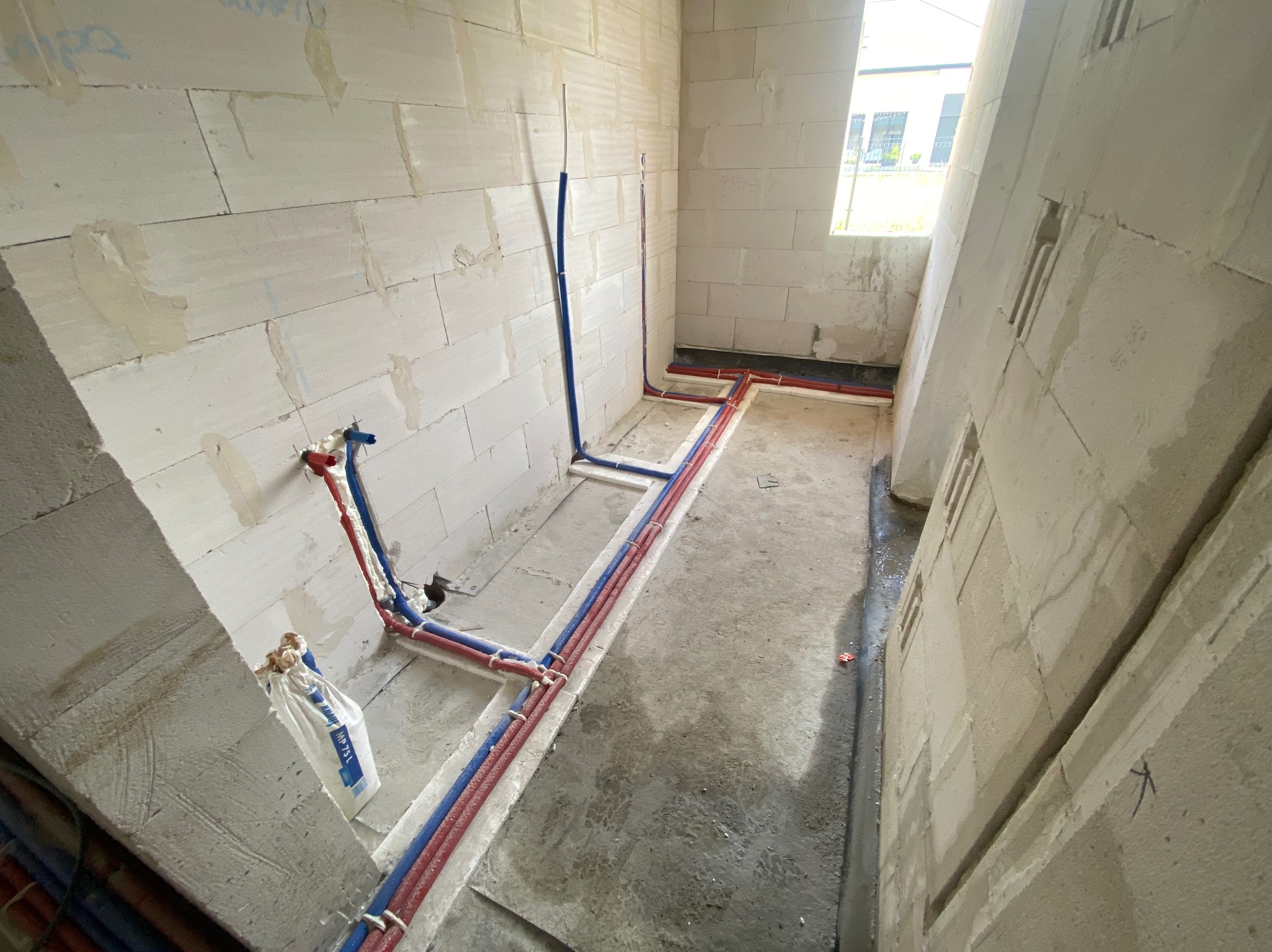 Wnętrze w trakcie instalacji hydraulicznej i ogrzewania podłogowego, widoczne rury czerwone i niebieskie prowadzone w bruzdach w ścianach z betonu komórkowego oraz w wylewce podłogowej, worek...