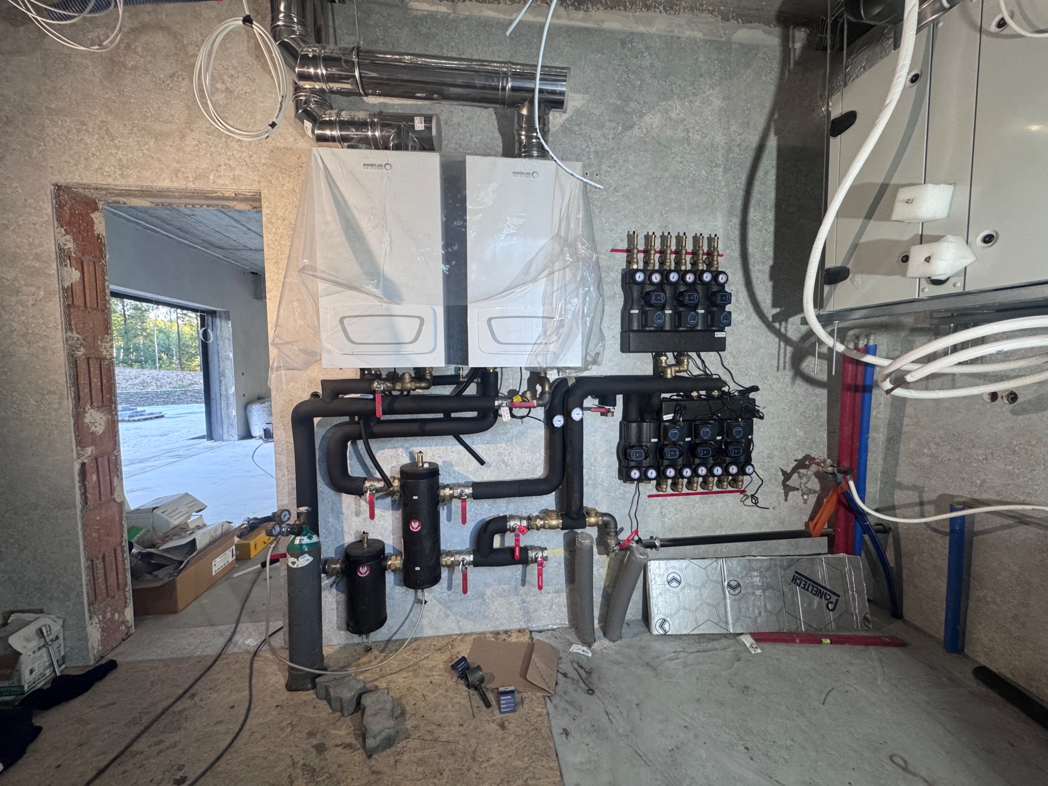 Dwa kotły grzewcze, czarne rury instalacji CO i rozdzielacze na surowej ścianie. Widoczne elementy instalacji i narzędzia. Stan surowy, przed wykończeniem.
