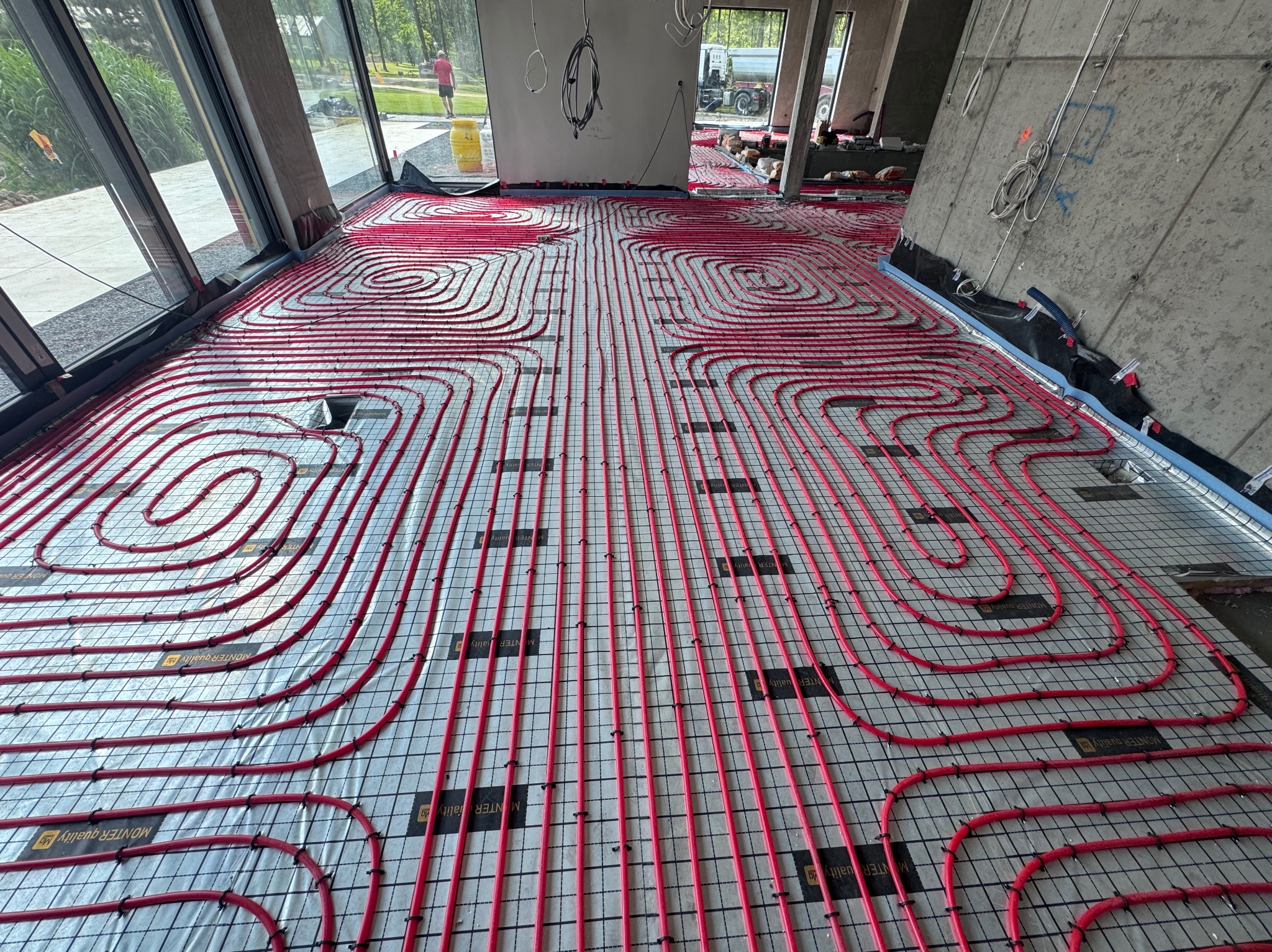 Czerwone rury ogrzewania podłogowego ułożone spiralnie na szarej macie w jasnym pomieszczeniu z betonową ścianą. Widoczne okna i osoba w oddali.