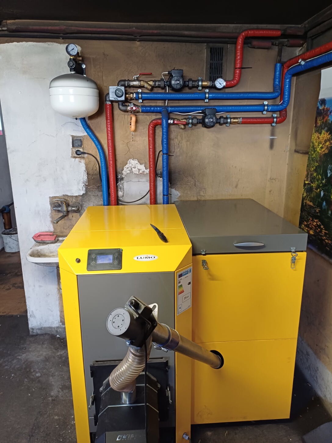Żółto-szary piec CO marki LUMO z podajnikiem, na tle ściany z instalacją grzewczą z rurami w izolacji czerwono-niebieskiej. Widoczna umywalka i narzędzia.