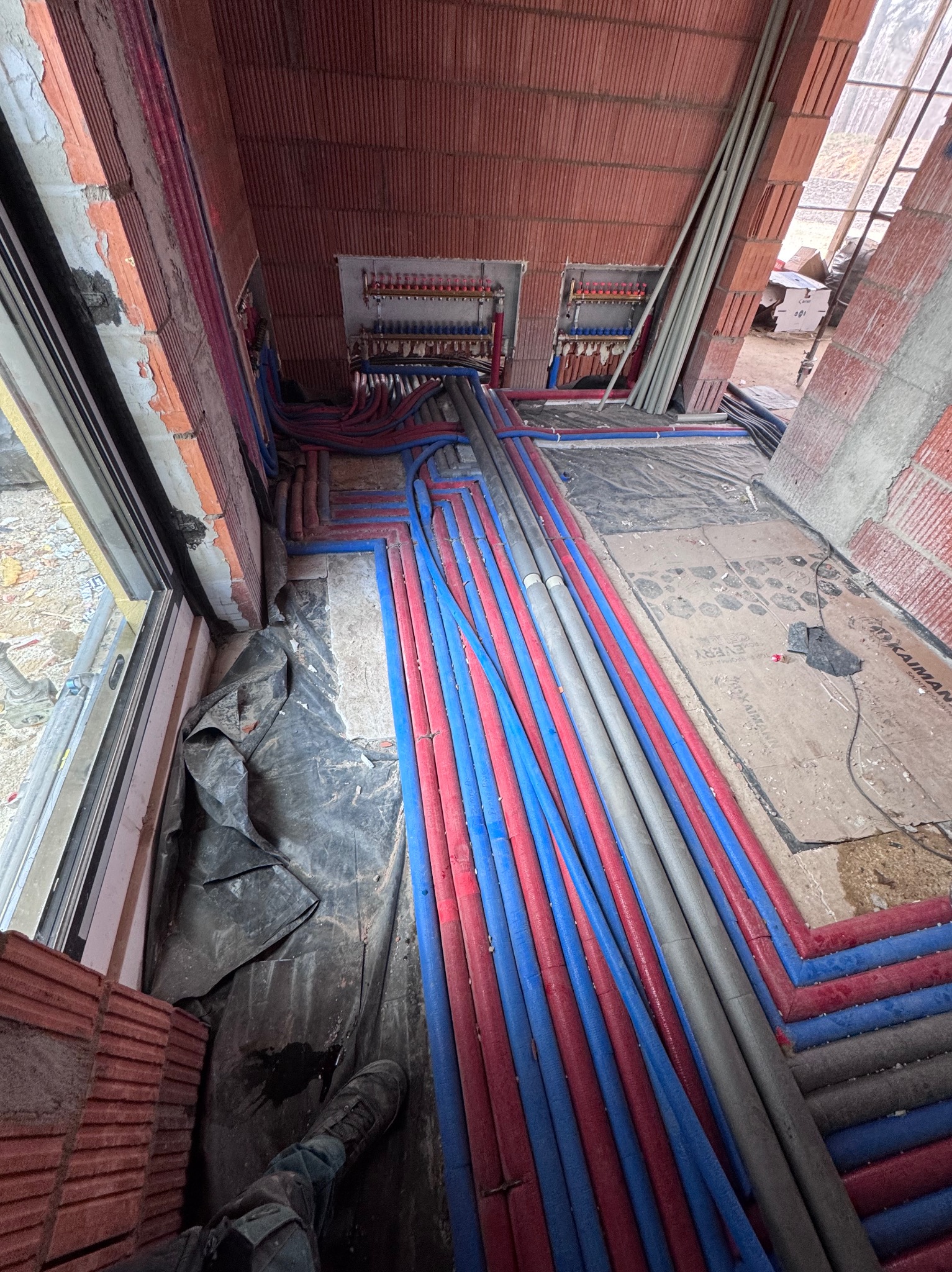 Instalacja ogrzewania podłogowego w trakcie montażu: rury grzewcze ułożone na podłodze w nowym budynku, widoczne rozdzielacze i fragmenty ścian z cegły.