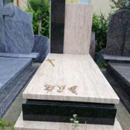 Jasny, polerowany pomnik z różowego granitu z czarnymi akcentami, ozdobiony metalowym krzyżem i trzema figurkami aniołów, ustawiony na zewnątrz wśród innych nagrobków.