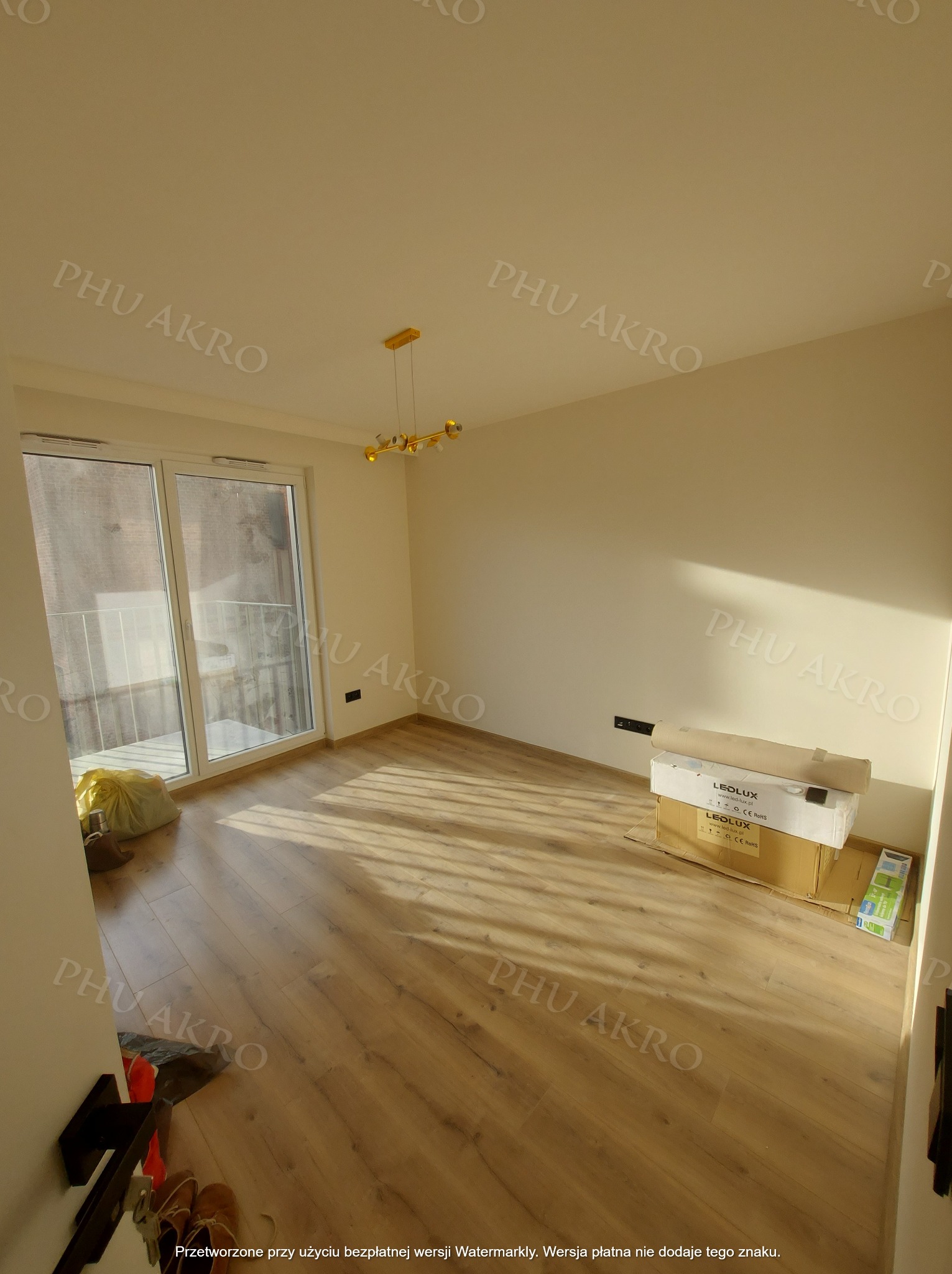 Jasne, puste pomieszczenie po remoncie z drewnianą podłogą, balkonem i złotą lampą. Kartony z oświetleniem LED czekają na montaż. Widok na zewnątrz przez balkon.