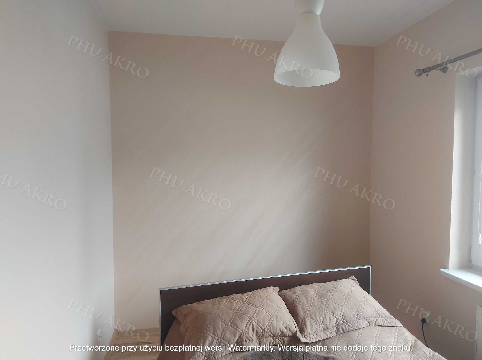 Pokój po odświeżeniu: ściany w odcieniach beżu, widoczne łóżko z brązową ramą i poduszkami. Lampa sufitowa w centralnej części kadru. Neutralne, ciepłe oświetlenie.