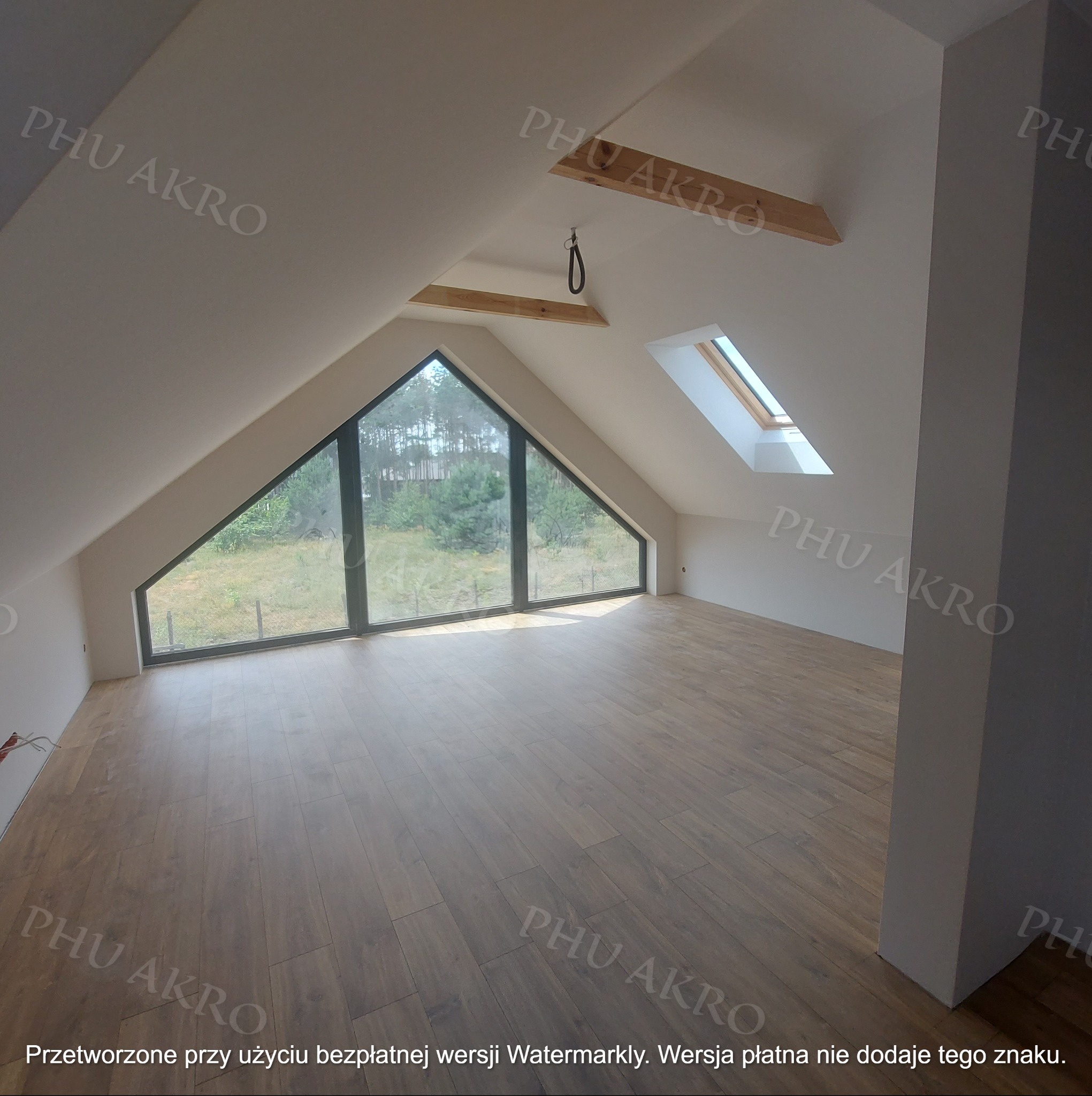 Jasne poddasze po remoncie z drewnianą podłogą i dużym oknem trójkątnym. Widoczne drewniane belki stropowe i okno dachowe. Ściany pomalowane na biało.