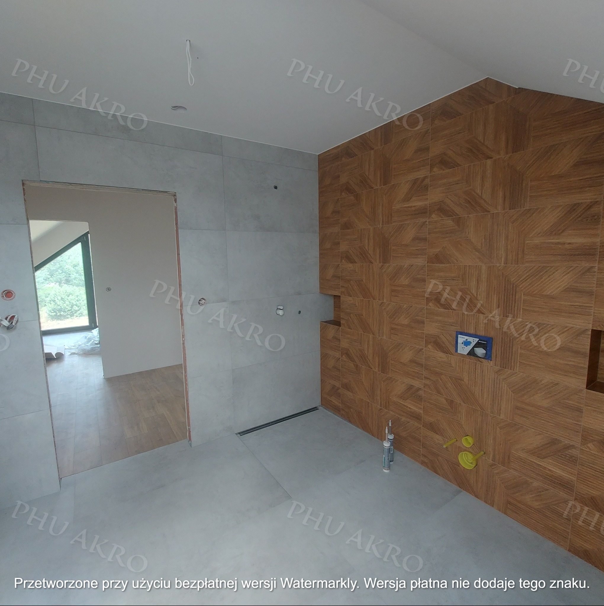 Wnętrze łazienki w trakcie prac. Widoczne płytki imitujące drewno na ścianie, szare płytki na podłodze i ścianach. Otwór drzwiowy prowadzi do innego pomieszczenia.