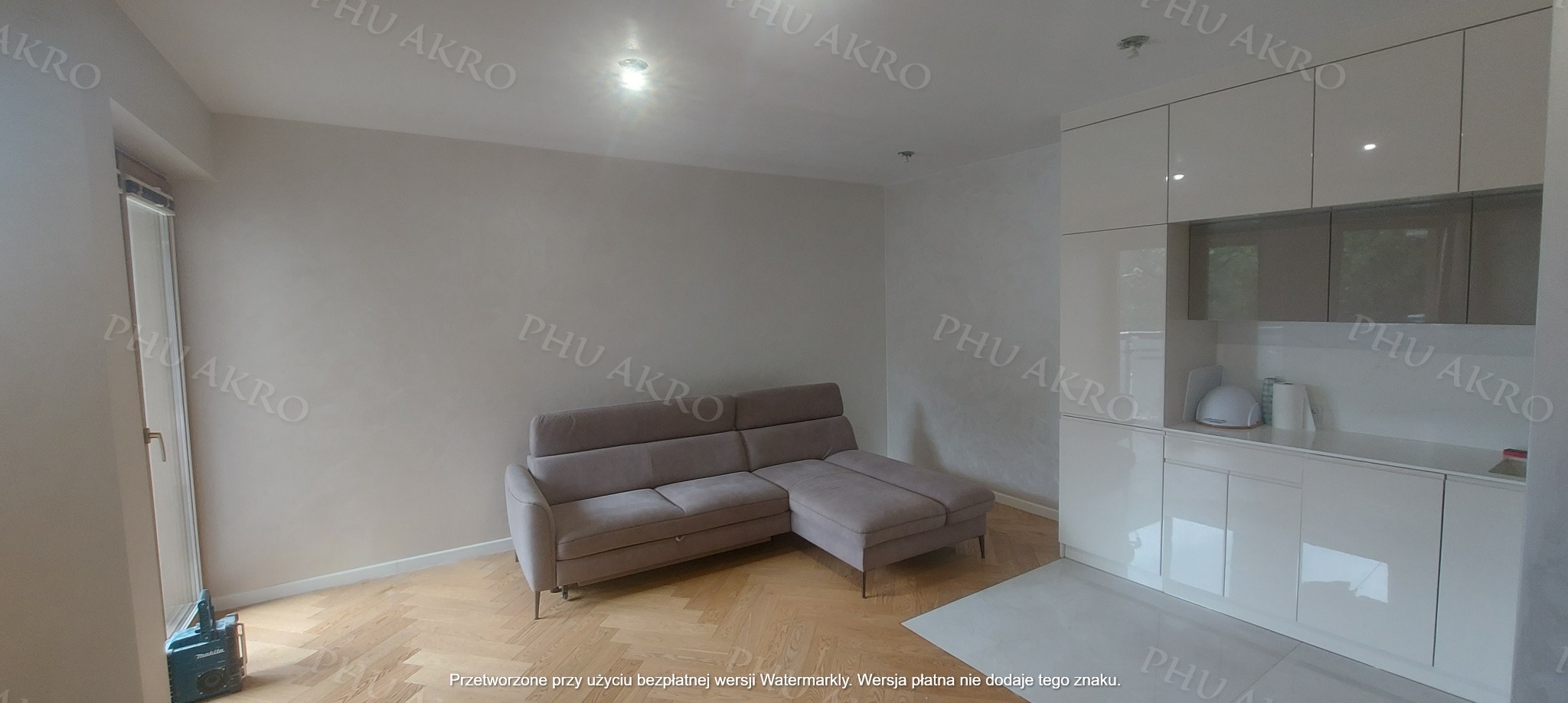 Jasne wnętrze po remoncie: szary narożnik, parkiet w jodełkę, biała zabudowa kuchenna z połyskiem. Widoczne narzędzie przy oknie. Minimalistyczny design.