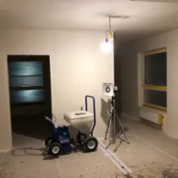 Wnętrze po nałożeniu gładzi, widoczny agregat malarski, lampa na statywie, okno zabezpieczone folią. Przygotowanie do dalszych prac.