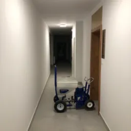 Korytarz po malowaniu natryskowym, widoczny agregat malarski Texspray. Świeżo pomalowane ściany w odcieniach bieli. Podłoga wyłożona płytkami w kolorze szarym.