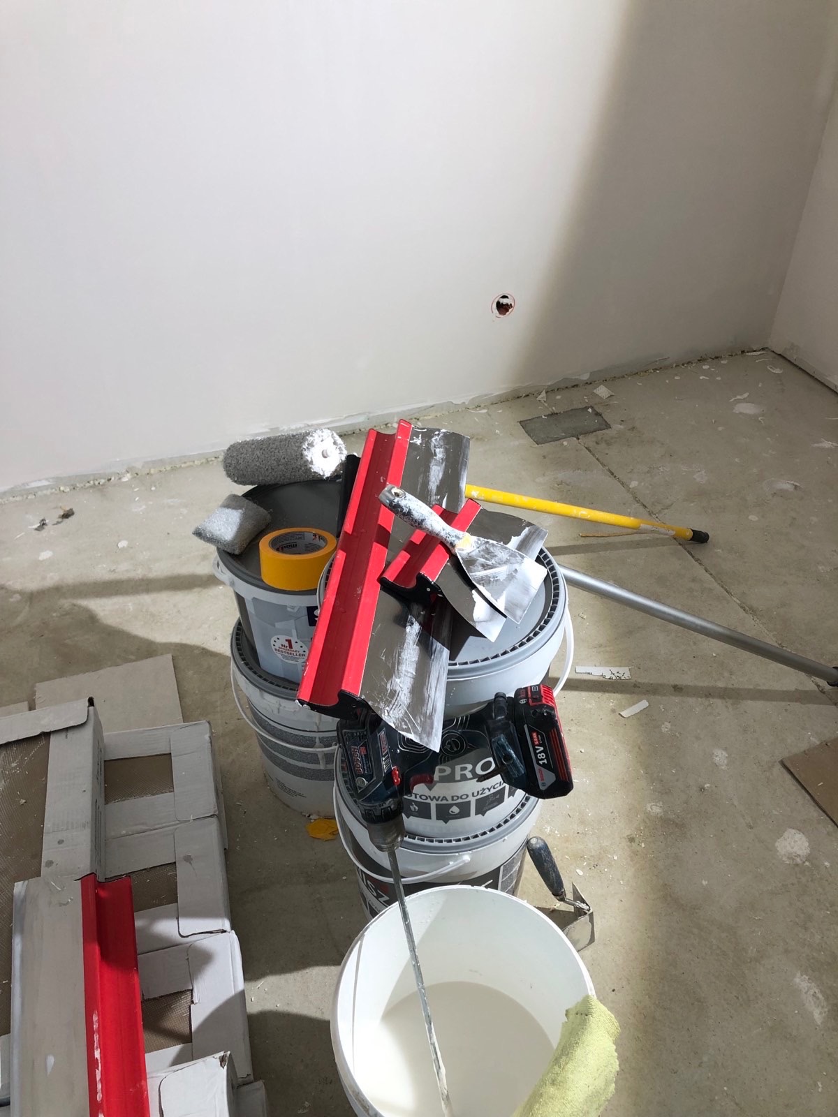 Wnętrze w trakcie prac wykończeniowych: wiadro z farbą, szpachelki, wałek malarski i mieszadło na tle gładkiej, pomalowanej ściany i surowej podłogi.
