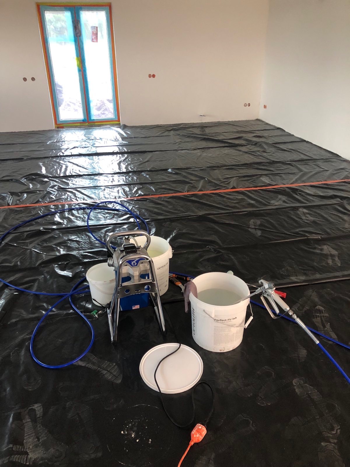 Przygotowanie do natryskowego malowania ścian: podłoga zabezpieczona folią, widoczny agregat malarski, pistolet natryskowy i wiadra z farbą w pomieszczeniu.