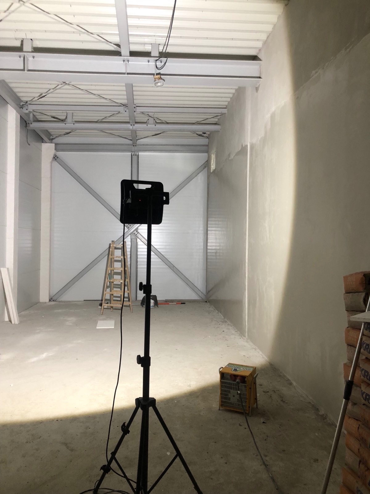 Wnętrze hali w trakcie prac wykończeniowych. Widoczna ściana z nałożoną warstwą gładzi, oświetlenie robocze na statywie i drabina oparta o ścianę. Hala z metalową konstrukcją.