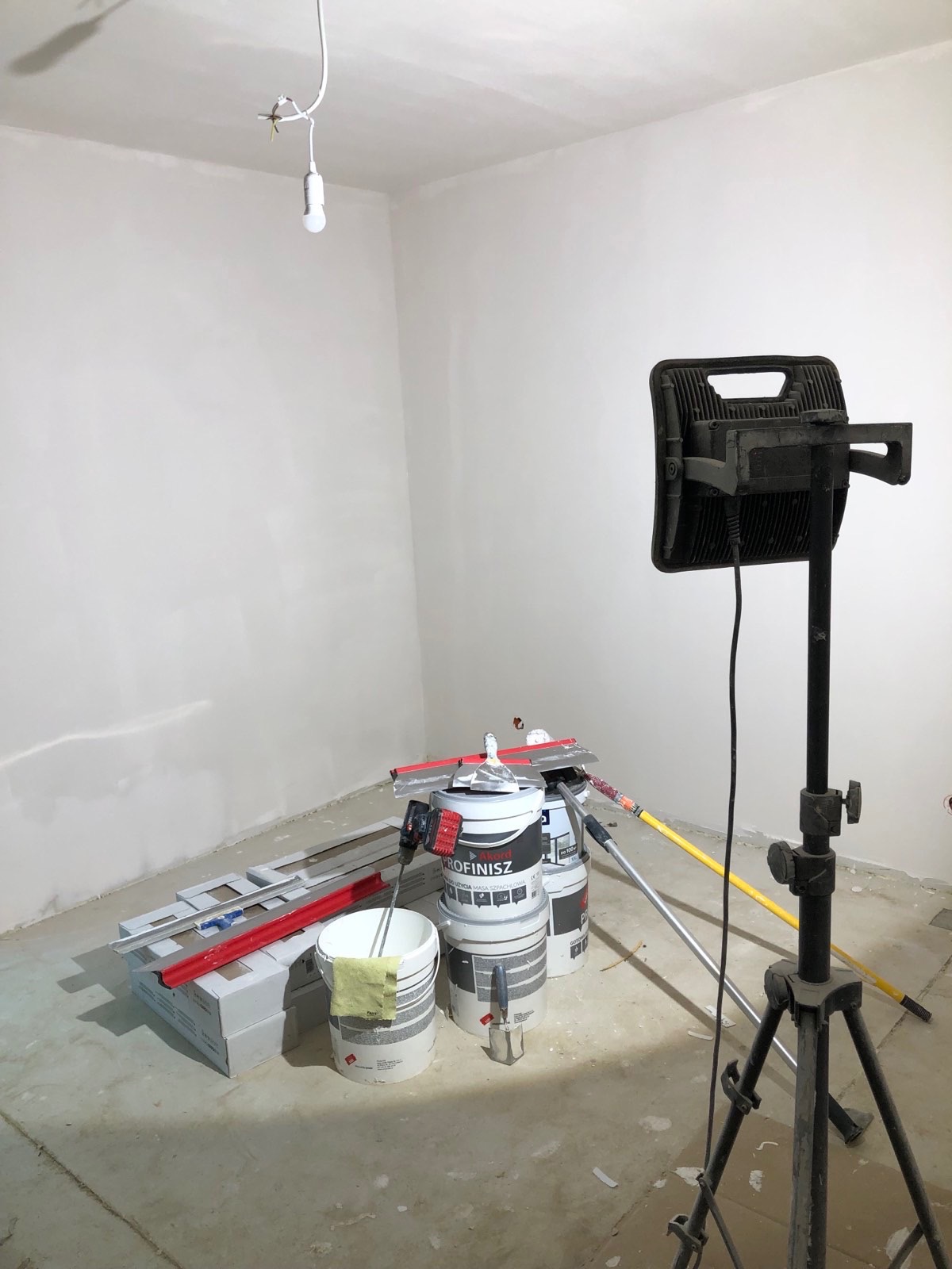 Ściany po nałożeniu gładzi, widoczne narzędzia i wiadra z masą szpachlową oraz lampa oświetleniowa na trójnogu. Przygotowanie do dalszych prac wykończeniowych.