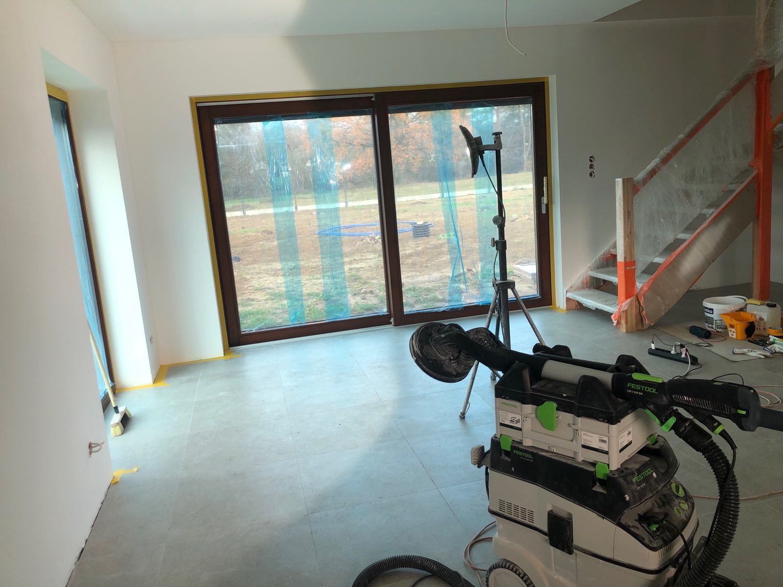 Wnętrze w trakcie prac: szlifierka do gładzi na podłodze z płytek, okna zabezpieczone folią, przygotowane ściany, schody w tle.