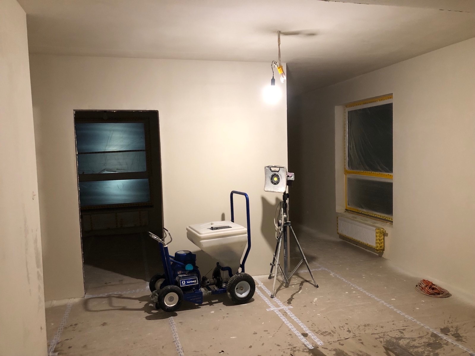 Wnętrze po nałożeniu gładzi, widoczny agregat malarski, lampa na statywie, okno zabezpieczone folią. Przygotowanie do dalszych prac.