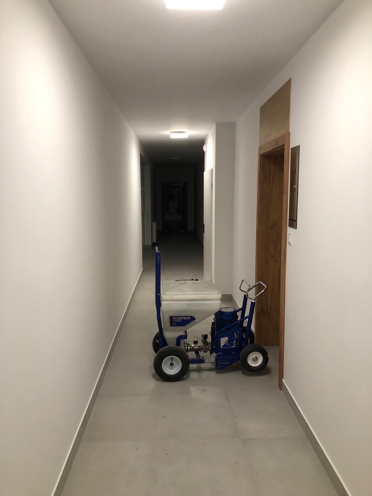 Korytarz po malowaniu natryskowym, widoczny agregat malarski Texspray. Świeżo pomalowane ściany w odcieniach bieli. Podłoga wyłożona płytkami w kolorze szarym.