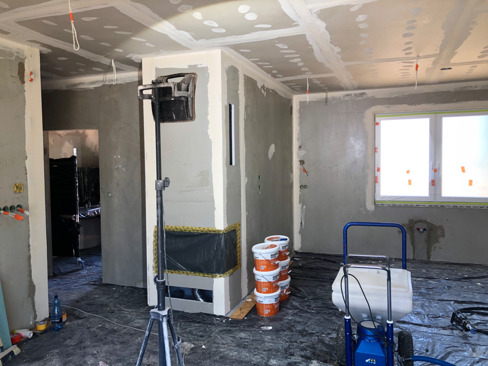 Wnętrze w trakcie prac wykończeniowych: ściany po szpachlowaniu, widoczny agregat malarski, wiaderka z masą szpachlową i lampa budowlana na trójnogu.
