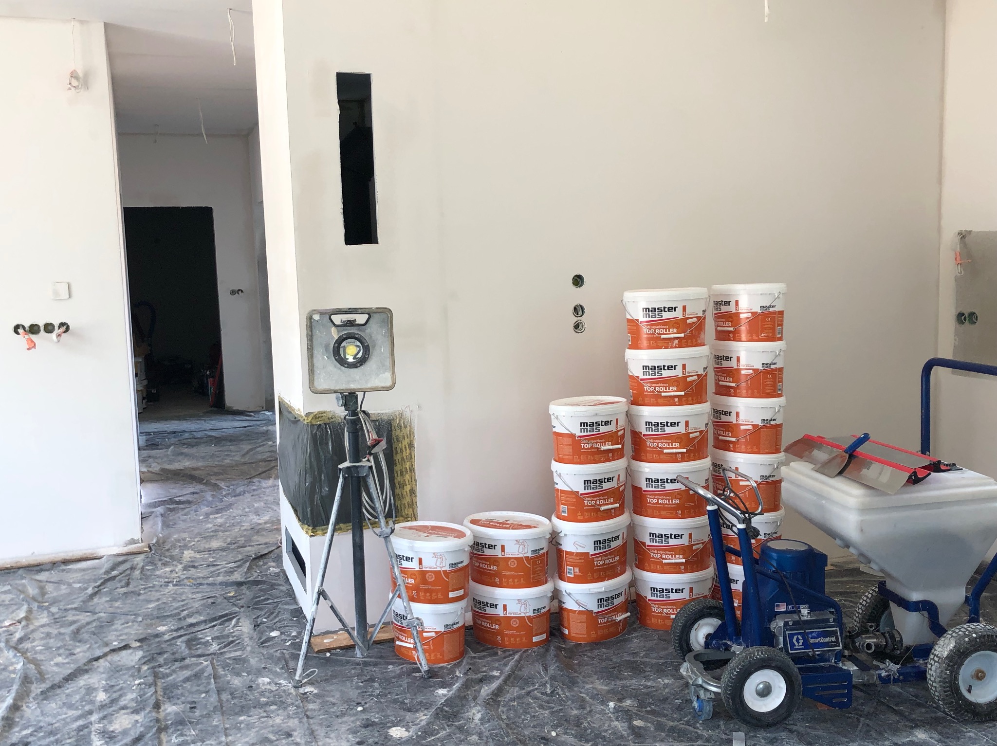 Wnętrze w trakcie prac: lampa budowlana, wiaderka z farbą Master Mas Top Roller i agregat malarski na folii ochronnej. Widoczny otwór w ścianie.