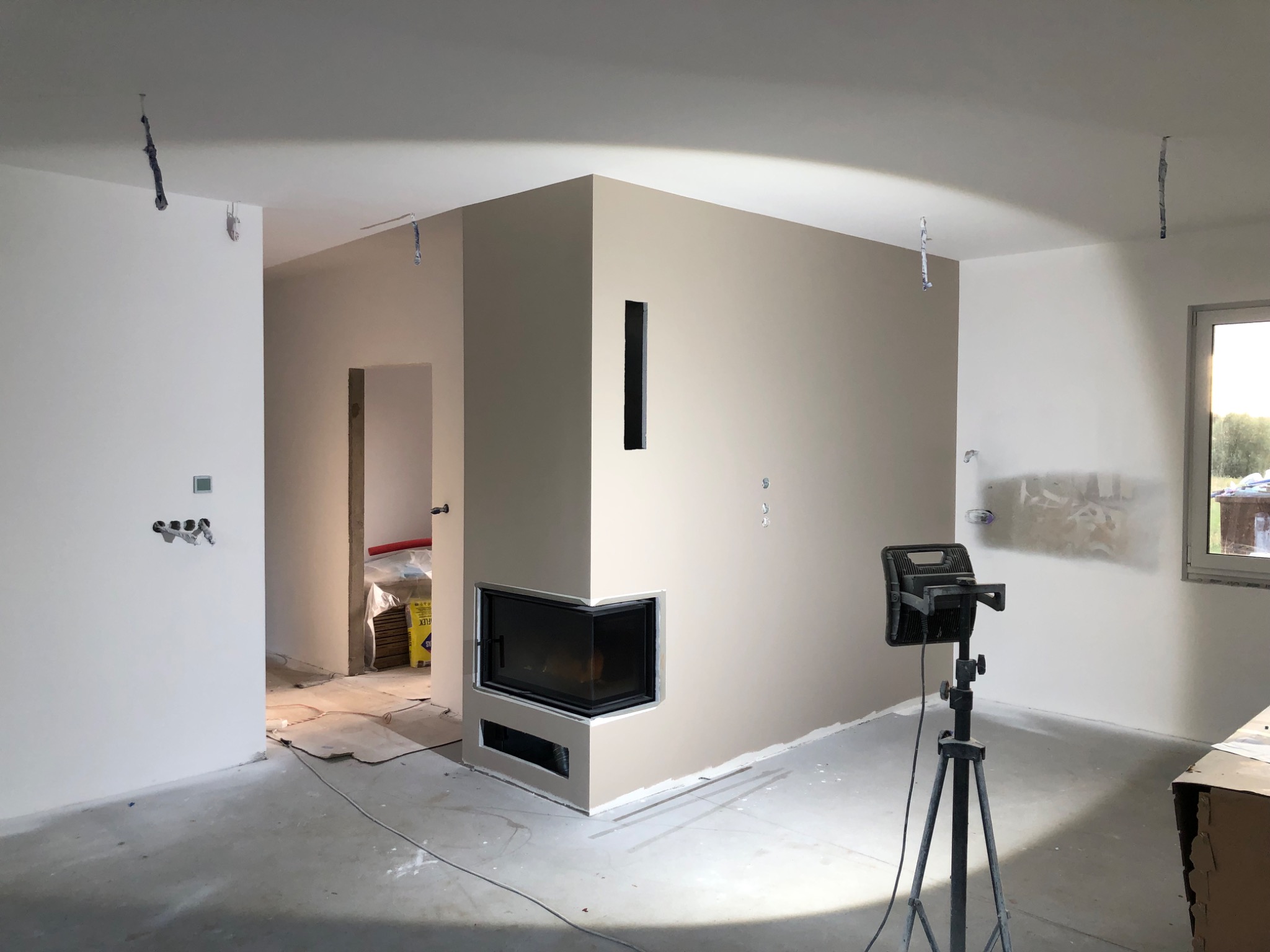 Wnętrze w trakcie prac wykończeniowych: ściany w dwóch odcieniach, zamontowany kominek narożny, widoczne kable i lampa na statywie. Częściowo wykończone pomieszczenie.