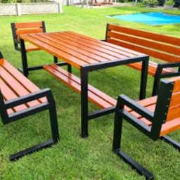 Zestaw mebli ogrodowych: stół z blatem z desek w odcieniach brązu oraz cztery ławki z oparciami, na zielonym trawniku, w tle ogrodzenie i roślinność.