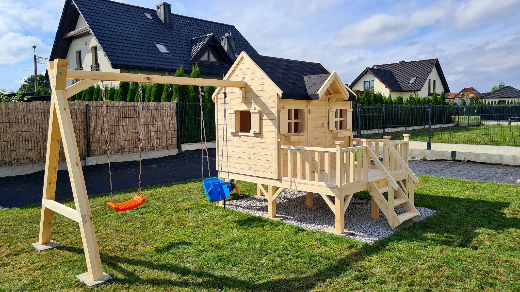 Drewniany domek na podwyższeniu z małym tarasem i huśtawką na tle zadbanego trawnika i domów jednorodzinnych.