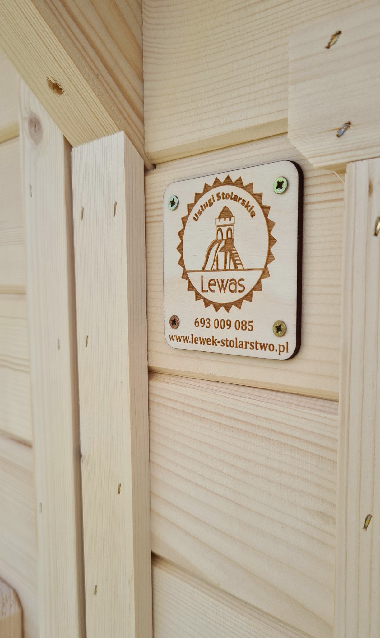 Drewniana tabliczka z logo firmy stolarskiej Lewas, przymocowana do jasnej, drewnianej konstrukcji, prawdopodobnie elementu placu zabaw.