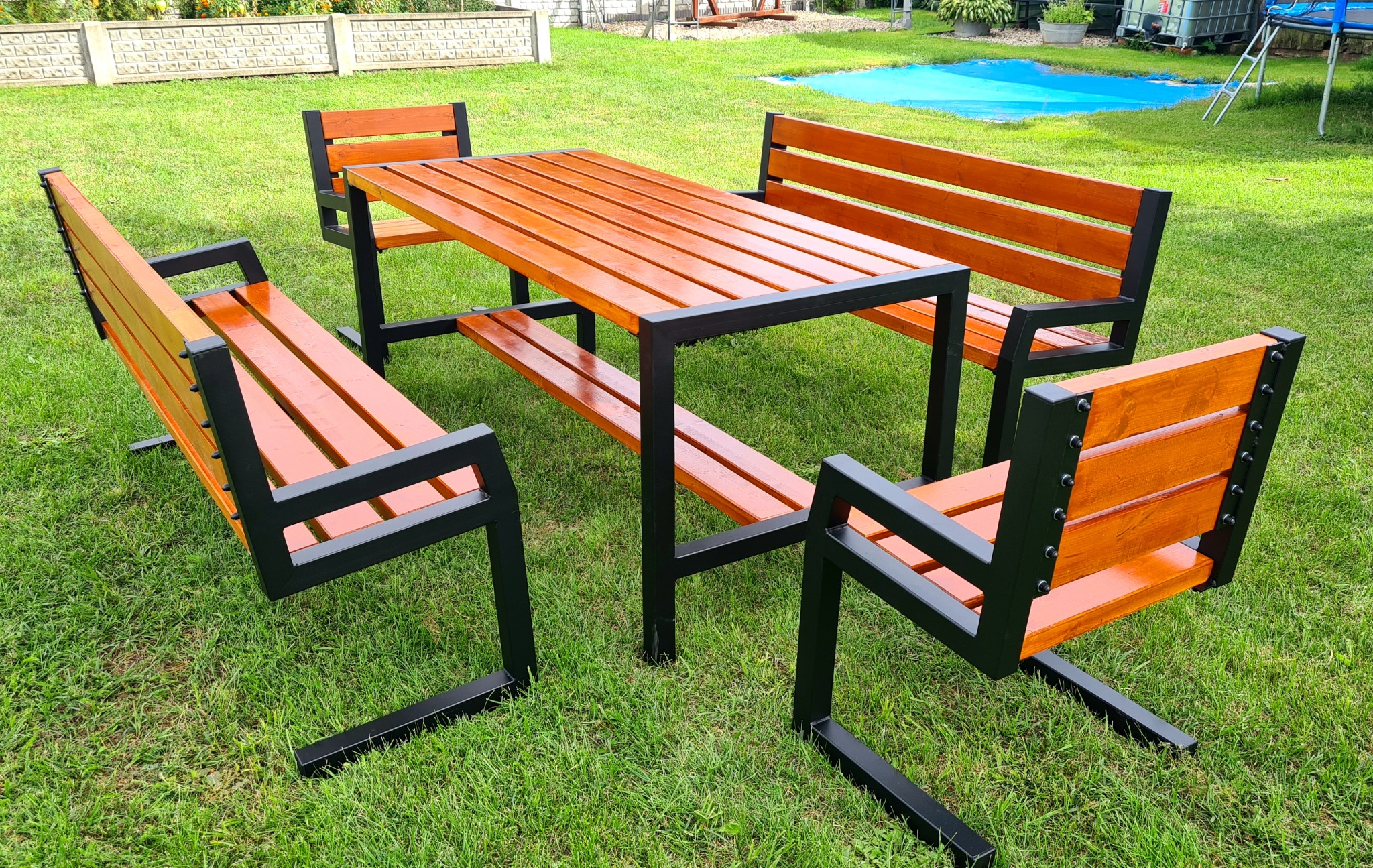 Zestaw mebli ogrodowych: stół z blatem z desek w odcieniach brązu oraz cztery ławki z oparciami, na zielonym trawniku, w tle ogrodzenie i roślinność.