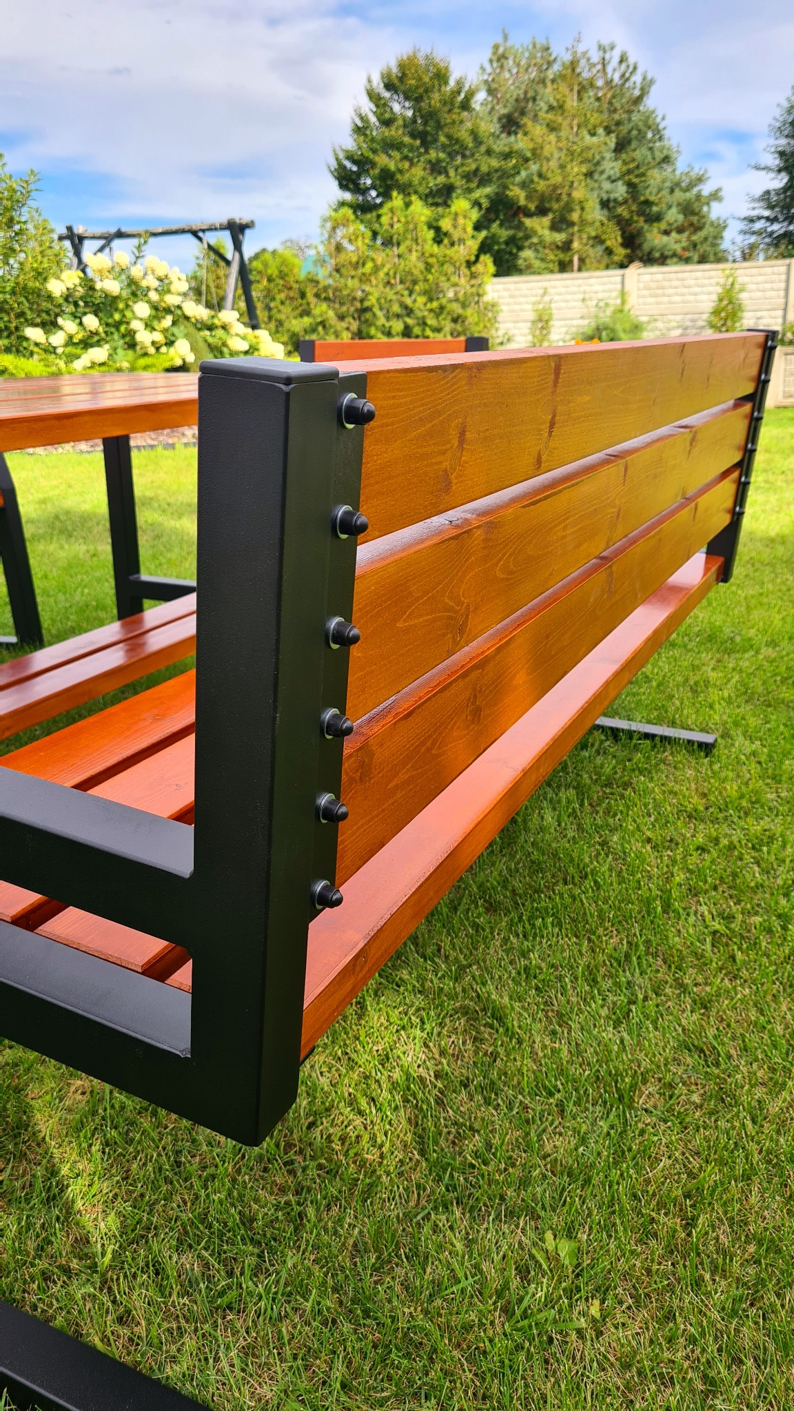 Fragment drewnianej ławki ogrodowej z czarną, metalową ramą, widocznej na tle trawnika, z elementami placu zabaw i roślinności w tle.