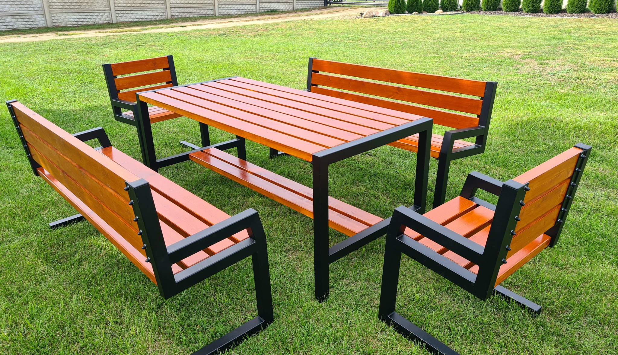 Zestaw mebli ogrodowych: stół z drewnianym blatem w kolorze miodowym i czarną metalową ramą oraz cztery ławki z oparciami, ustawiony na trawniku przed betonowym ogrodzeniem.