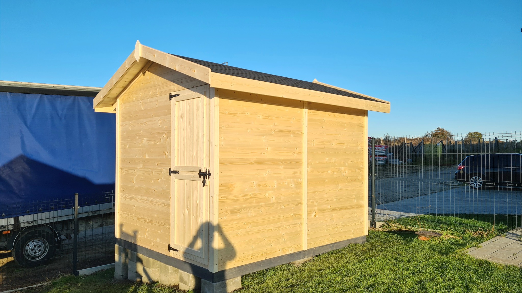 Drewniana altana ogrodowa z czarnym dachem i okuciami, ustawiona na betonowych bloczkach, widoczna na tle trawnika i częściowo zasłoniętej plandeką ciężarówki.