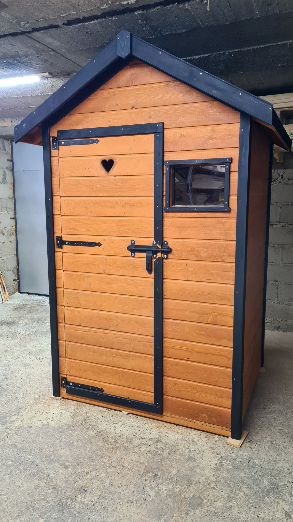 Drewniany domek dziecięcy z małym oknem, drzwiami z wyciętym sercem i czarnymi elementami wykończeniowymi, stojący na betonowej posadzce w szarym pomieszczeniu.
